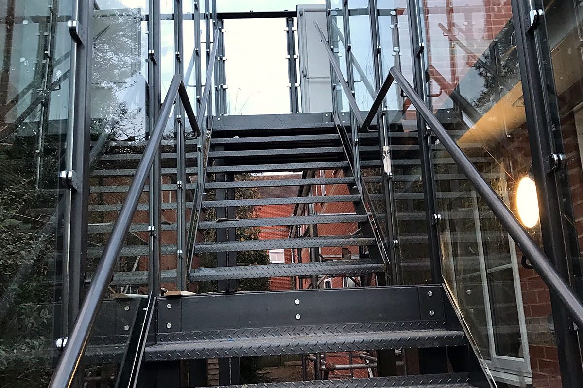 Black steel straight external stair with chequer plate treads inside a glass enclosure beside a brick building
