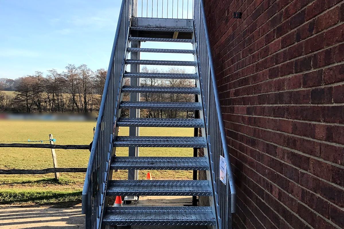 Galvanised steel staircase with chequer-plate treads rising beside a brick wall to a first-floor balcony
