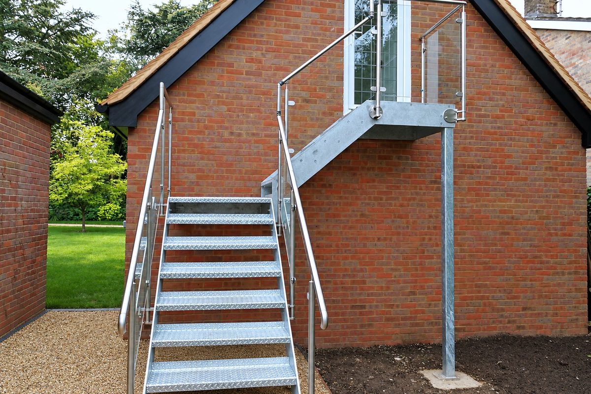 Galvanised steel stair with chequer plate treads up to a first-floor door, with a glass balustrade on the landing