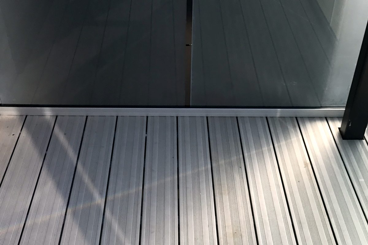 Two clear glass balustrade panels standing in a slim silver base channel on grey decking, beside a dark grey post