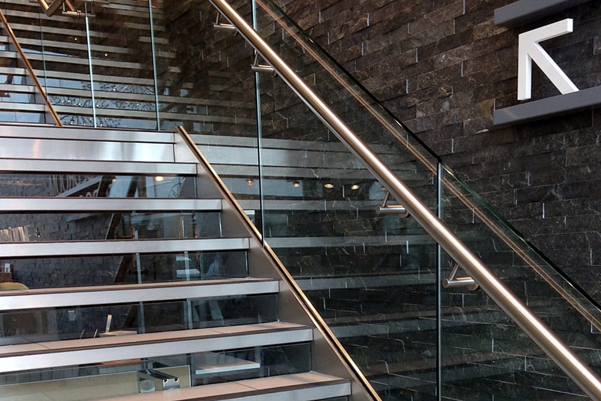 Stainless steel staircase with a frameless glass balustrade and a bronze-coloured handrail against a dark tiled wall
